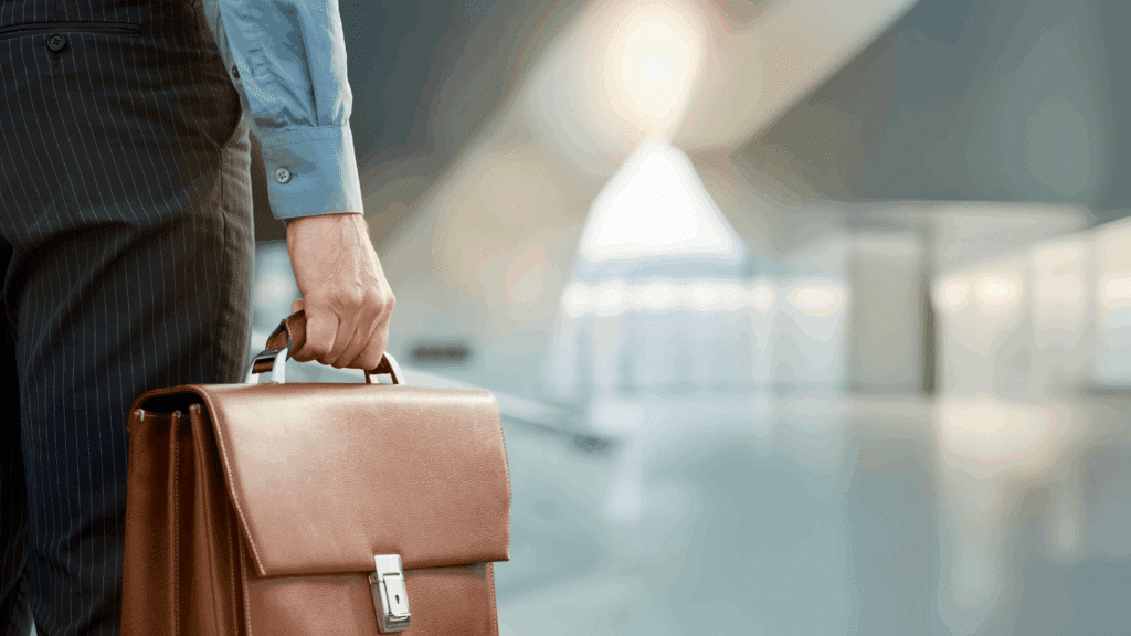 Close-up of a man in formal attire holding a leather briefcase, standing in a modern hallway with a blurred office background