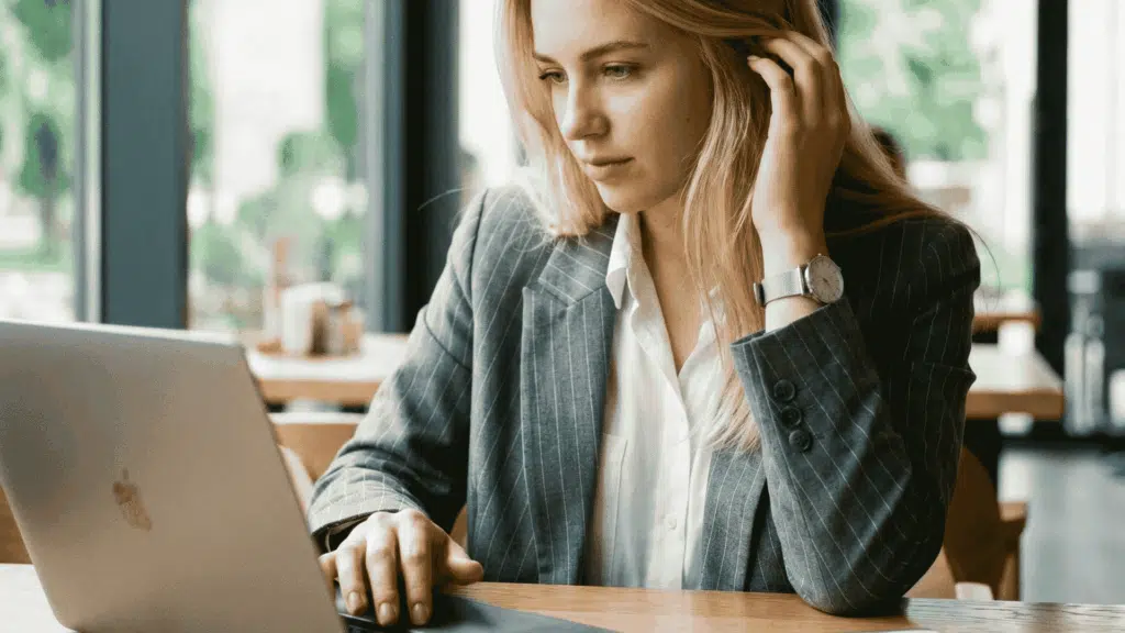 A blonde woman in a pinstripe blazer works on a laptop at a cafe table next to her smartphone