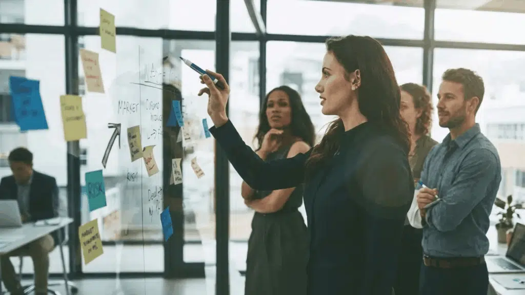 A shift leader guiding a team during a workplace meeting while presenting ideas on a glass board covered with notes and workflow plans.