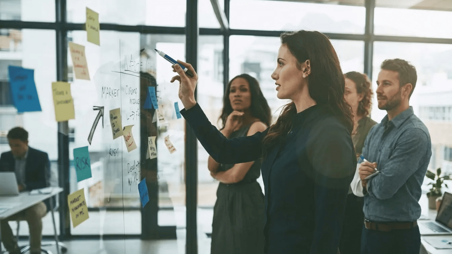 A shift leader guiding a team during a workplace meeting while presenting ideas on a glass board covered with notes and workflow plans.