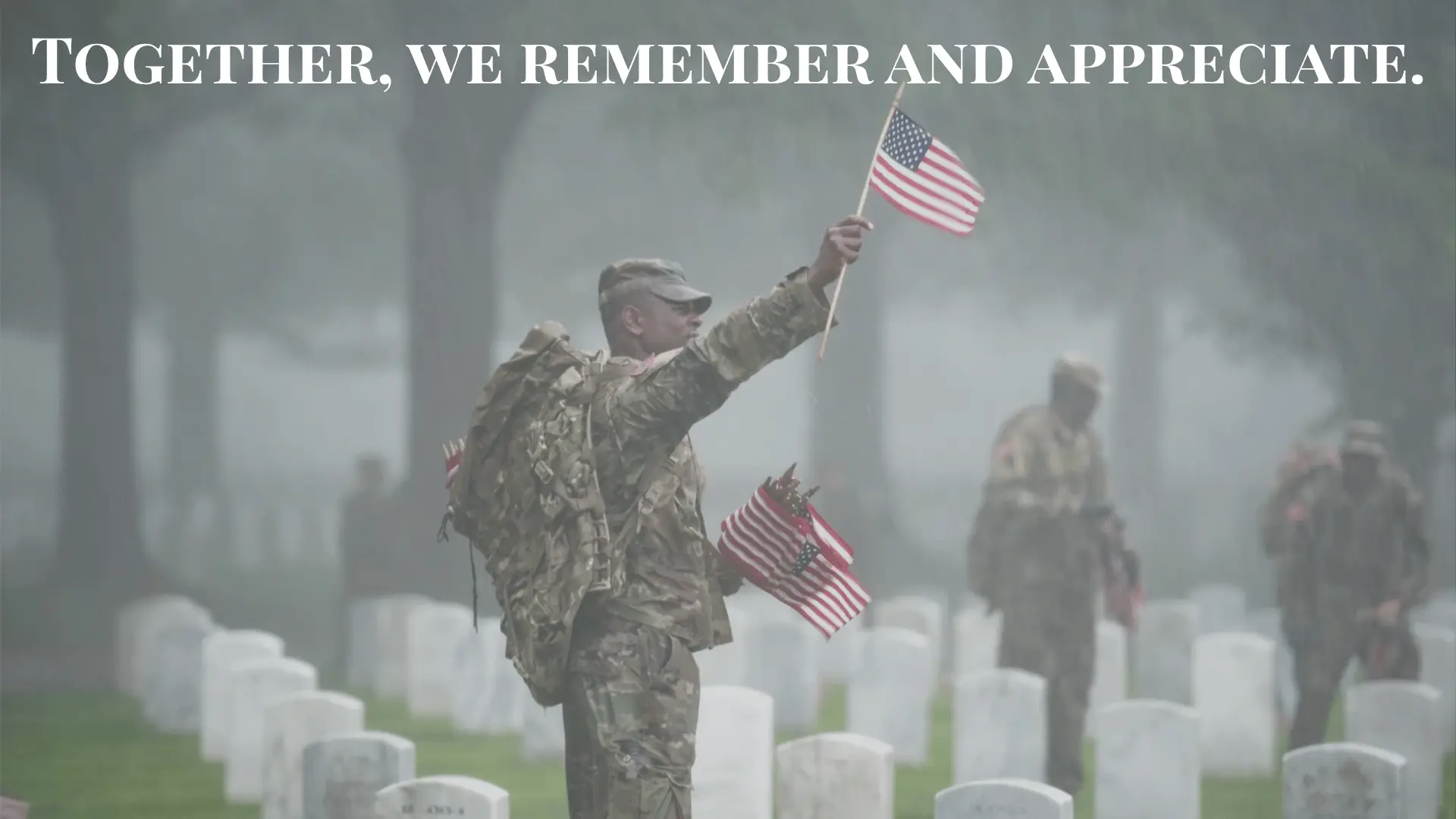 Soldier holding an American flag in a cemetery, with fellow soldiers placing flags on gravestones in the rain.