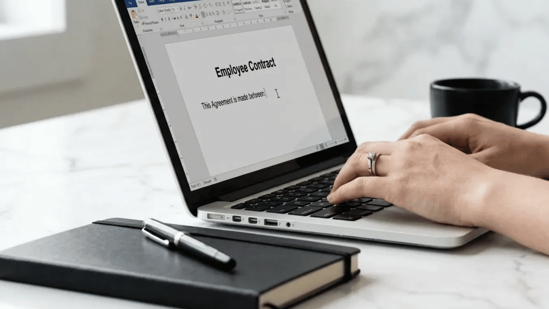 Person typing an employee contract on a laptop, with a notebook, pen, and coffee cup on a white desk.
