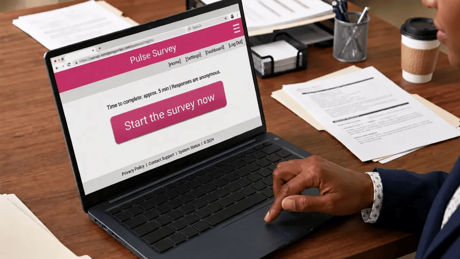 Person using a laptop at a desk to complete a pulse survey on a pulse survey software, with the screen showing a “Start the survey now” button, surrounded by documents and a coffee cup.
