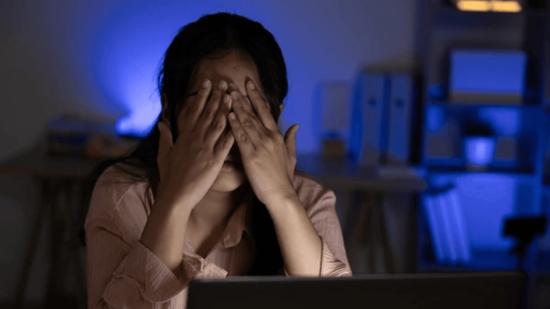 Woman covering her face with both hands while sitting in front of a laptop at night
