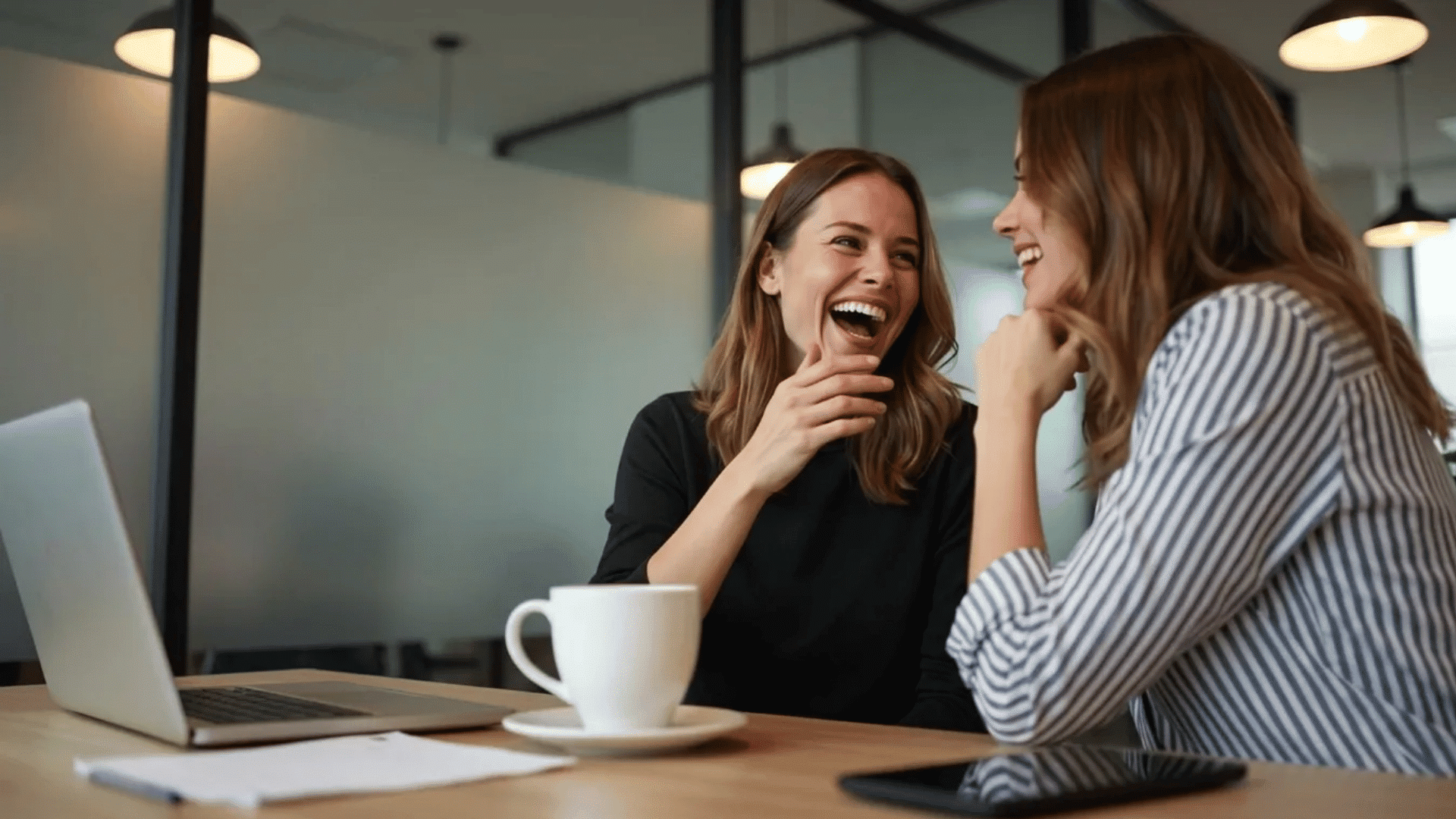 Two coworkers laughing together during a casual office conversation beside a laptop, coffee cup, and tablet.
