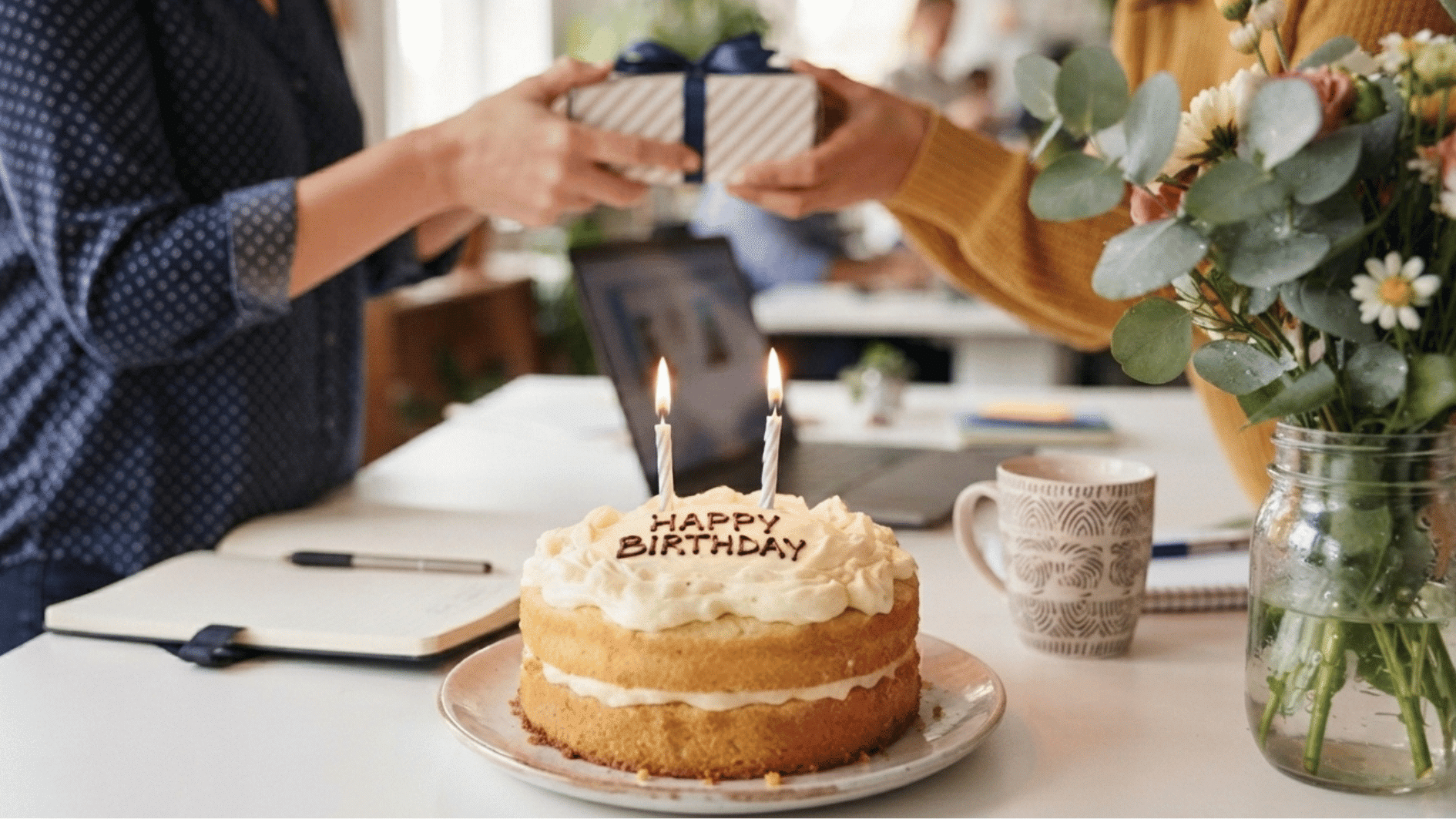 two hands giving and receiving birthday gift, with a birthday cake on desk.