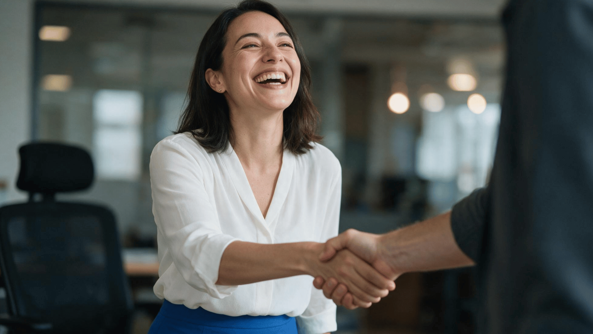 a smiling woman in professional attire enthusiastically shakes hands with a colleague in a modern office