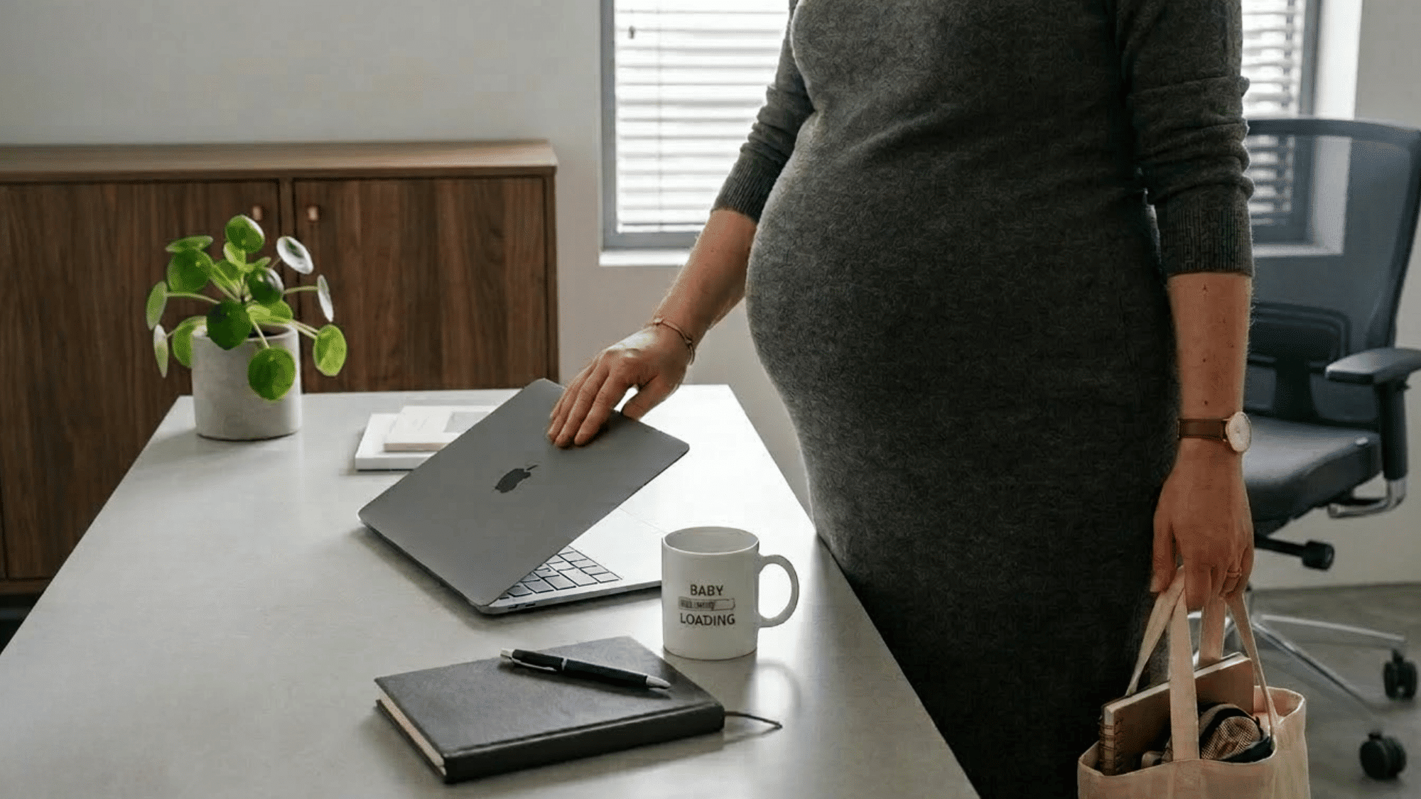 pregnant employee closing her laptop and leaving an office with a work bag beside a desk and baby loading mug